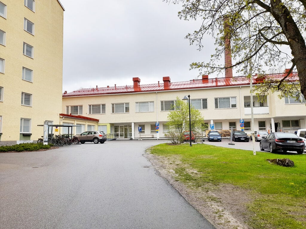 Kuva pihasta pilvisenä päivänä. Etualalla näkyy asfaltoitu piha-alue, jonka vasemmalla puolella on polkupyöräteline ja pyöriä sen alla. Taustalla on kaksikerroksinen vaalea rakennus, jossa on punainen peltikatto ja useita savupiippuja. Vasemmalle jää kuusikerroksinen vaalea kivitalo. Pihan oikealla puolella on pysäköityjä autoja ja viheralue, jossa kasvaa puu.