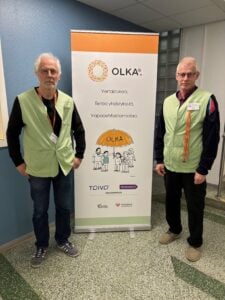Kaksi OLKA-vapaaehtoista miestä seisoo roll-up-julisteen vieressä sisätiloissa. Molemmat miehet käyttävät vaaleanvihreitä OLKA-liivejä ja nimikylttiä. Vasemmalla oleva mies on harmaahiuksinen, käyttää silmälaseja, mustaa paitaa ja farkkuja. Oikealla oleva mies on myös harmaahiuksinen ja silmälasipäinen, pukeutunut tummaan paitaan ja mustiin housuihin. Roll-upissa lukee: ”OLKA® – Vertaistukea. Tietoa yhdistyksistä. Vapaaehtoistoimintaa.” Julisteessa on piirretty kuva ihmisistä OLKA-sateenvarjon alla. Alhaalla näkyvät logot: TOIVO, Vertaistalo.fi, Etelä-Karjalan Yhdistykset ry ja Etelä-Karjalan hyvinvointialue.