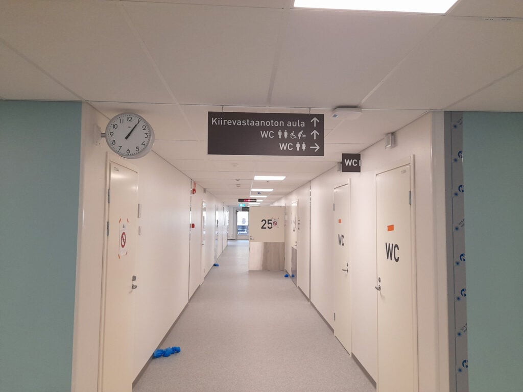 Pitkänmallinen vaalea käytävä, jonka molemmin puolin valkoisia ovia. Etualalla käytävän katossa suurikokoinen kellotaulu. Käytävän päässä häämöttää ulko-ovi.