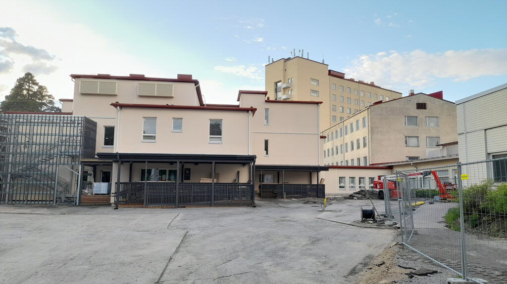 Panoraamakuva vaaleasta rakennuksesta, jonka edessä on harmaa hiekkainen piha-alue. Rakennuksessa on tiloja eri tasoissa. Rakennuksen sisäänkäynnin edessä on tummanharmaat katoksen aitaukset.