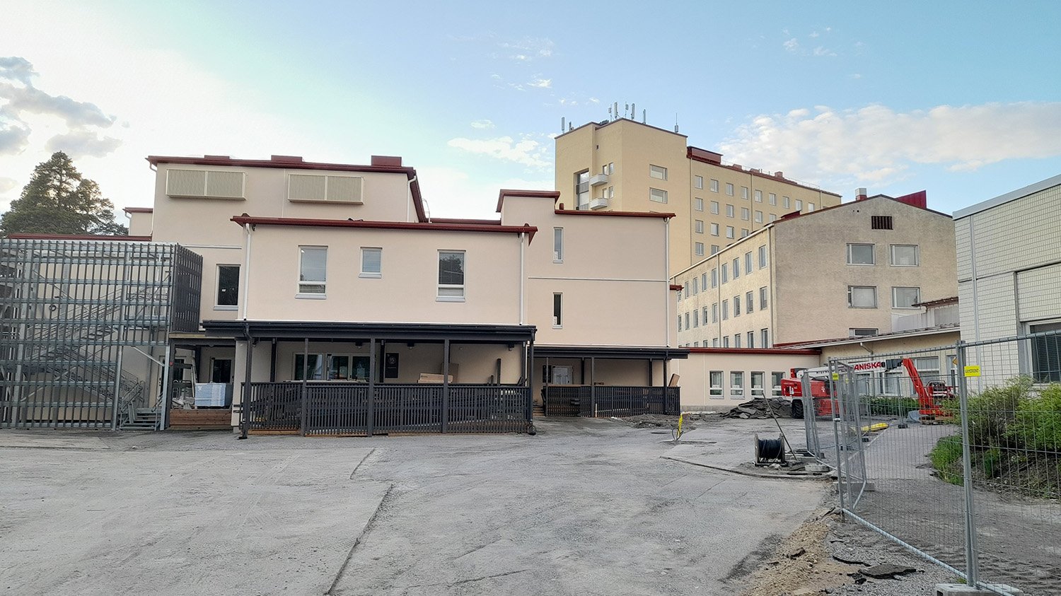Panoraamakuva vaaleasta rakennuksesta, jonka edessä on harmaa hiekkainen piha-alue. Rakennuksessa on tiloja eri tasoissa. Rakennuksen edessä on tummanharmaat