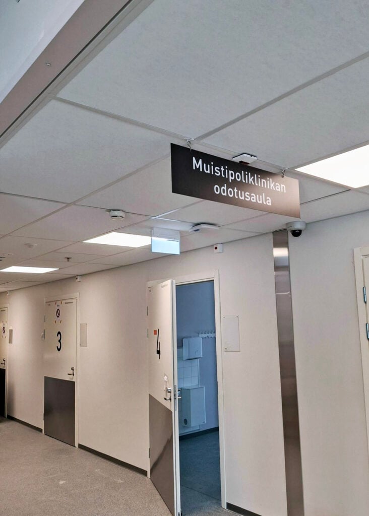 Vaalea käytävä, jossa yksi ovi osittain avoinna, muut kaksi ovea ovat kiinni. Käytävän yllä roikkuu kyltti, jossa lukee Muistipoliklinikka odotusaula.