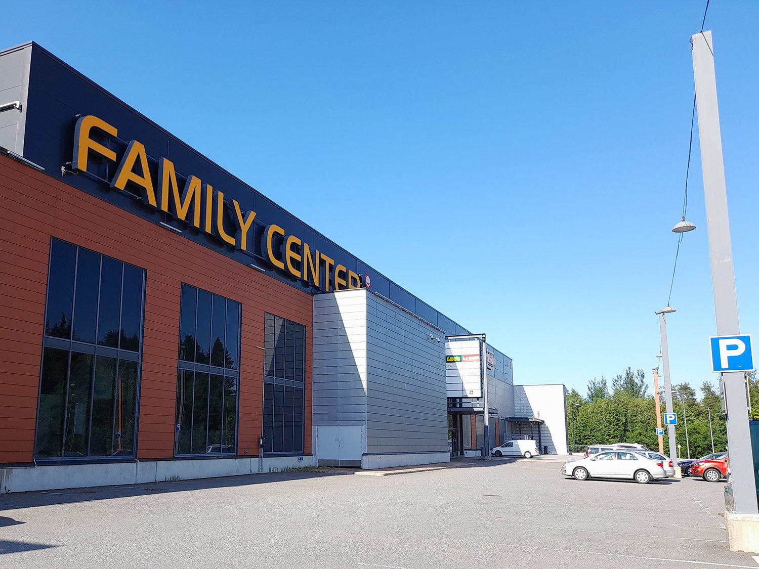 Kauppakeskus nimeltään "Family Center" aurinkoisana päivänä. Rakennus on moderni, kaksikerroksinen ja julkisivultaan punatiilen ja harmaan sävyinen. Etualalla on iso, tyhjä parkkialue ja muutamia pysäköityjä autoja.