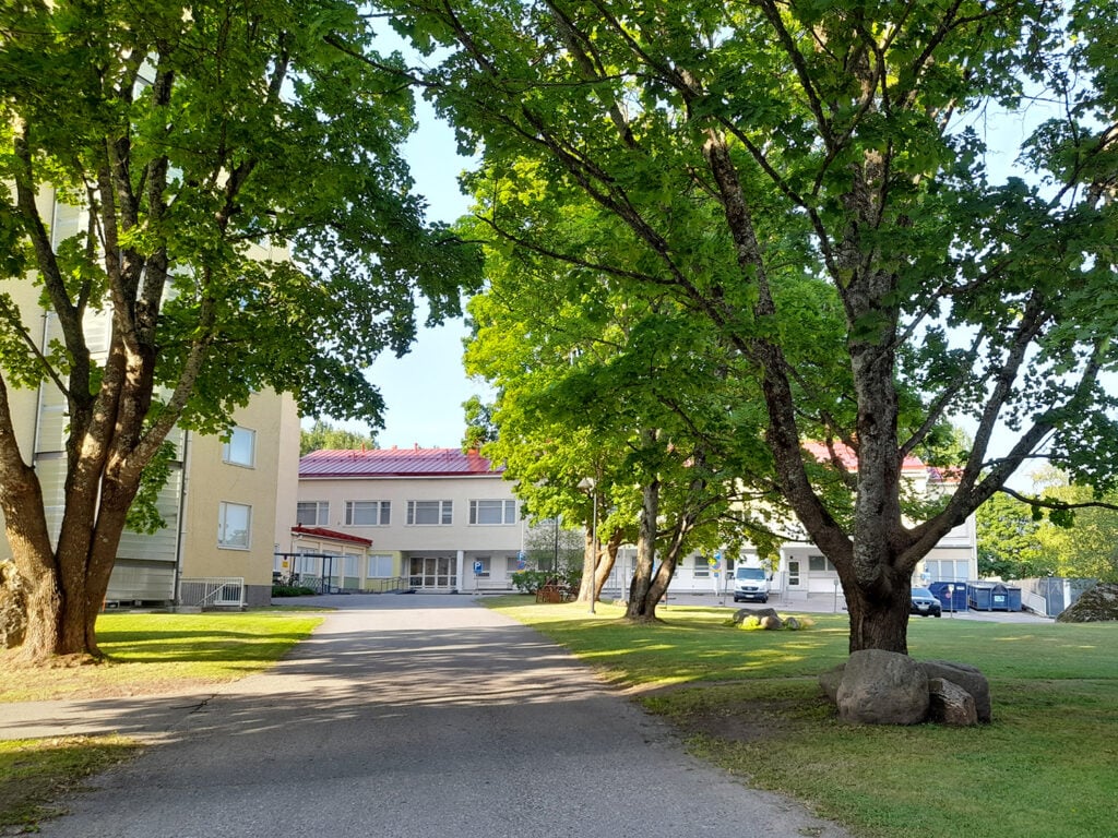 Puutarhamainen pihatie, jota reunustavat suuret vihreät puut. Taustalla näkyy vaalea rakennus punaisella katolla, ja oikealla on kiviryhmä nurmikolla.