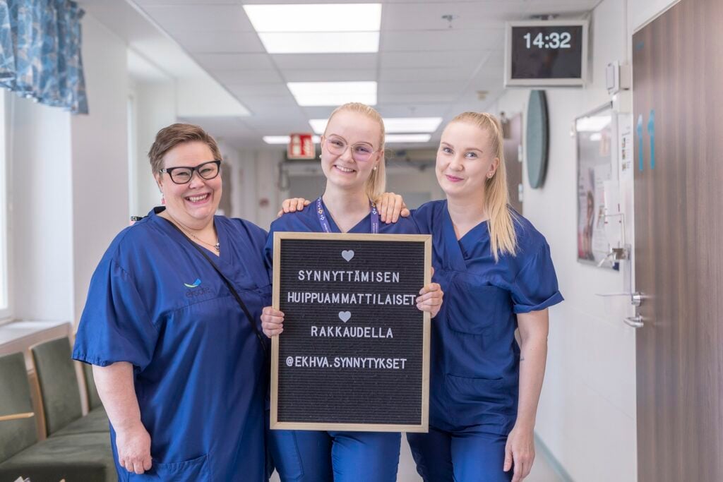 Kolme hymyilevää sairaanhoitajaa tai kätilöä seisoo sairaalan käytävällä sinisissä työasuissa. Keskimmäinen henkilö pitelee kylttiä, jossa lukee: "❤️ Synnyttämisen huippuammattilaiset ❤️ Rakkaudella @ekhva.synnytykset". Taustalla näkyy sairaalan käytävä, huoneiden ovia ja digitaalinen kellonäyttö, jossa lukee 14:32.