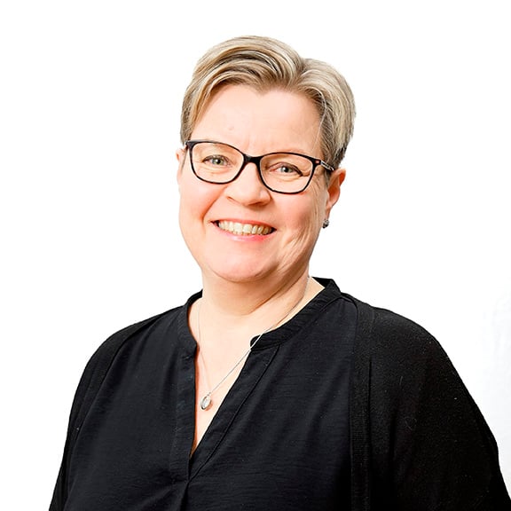 Vaalea lyhyttukkainen hymyilevä nainen, jolla tummasankaiset silmälasit. Päällään hänellä on musta pitkähihainen ja V-aukkoinen pusero.