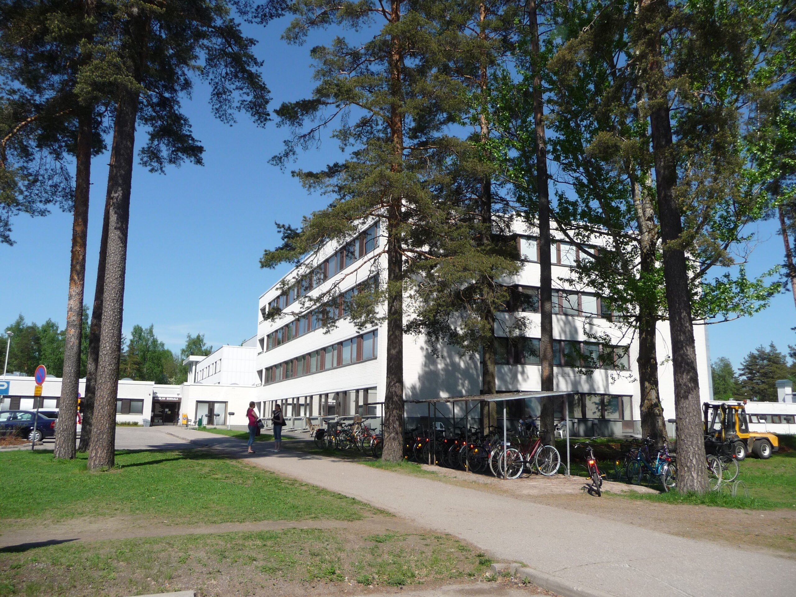 Vaalea rakennus useilla ikkunoilla, etualalla pyöräteline ja nurmikkoa. Rakennuksen edessä kulkee jalkakäytävä, ja ympärillä on runsaasti vihreitä puita kirkkaassa päivänvalossa.