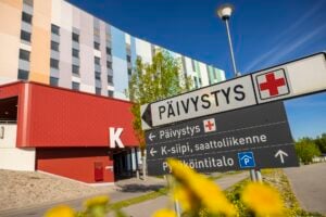 Sairaalan edustalla oleva kyltti, jossa lukee 'Päivystys' punaisen ristin symbolin kanssa. Taustalla näkyy monivärinen sairaalarakennus, punainen K-ovi ja sininen taivas. Etualalla on keltaisia kukkia.