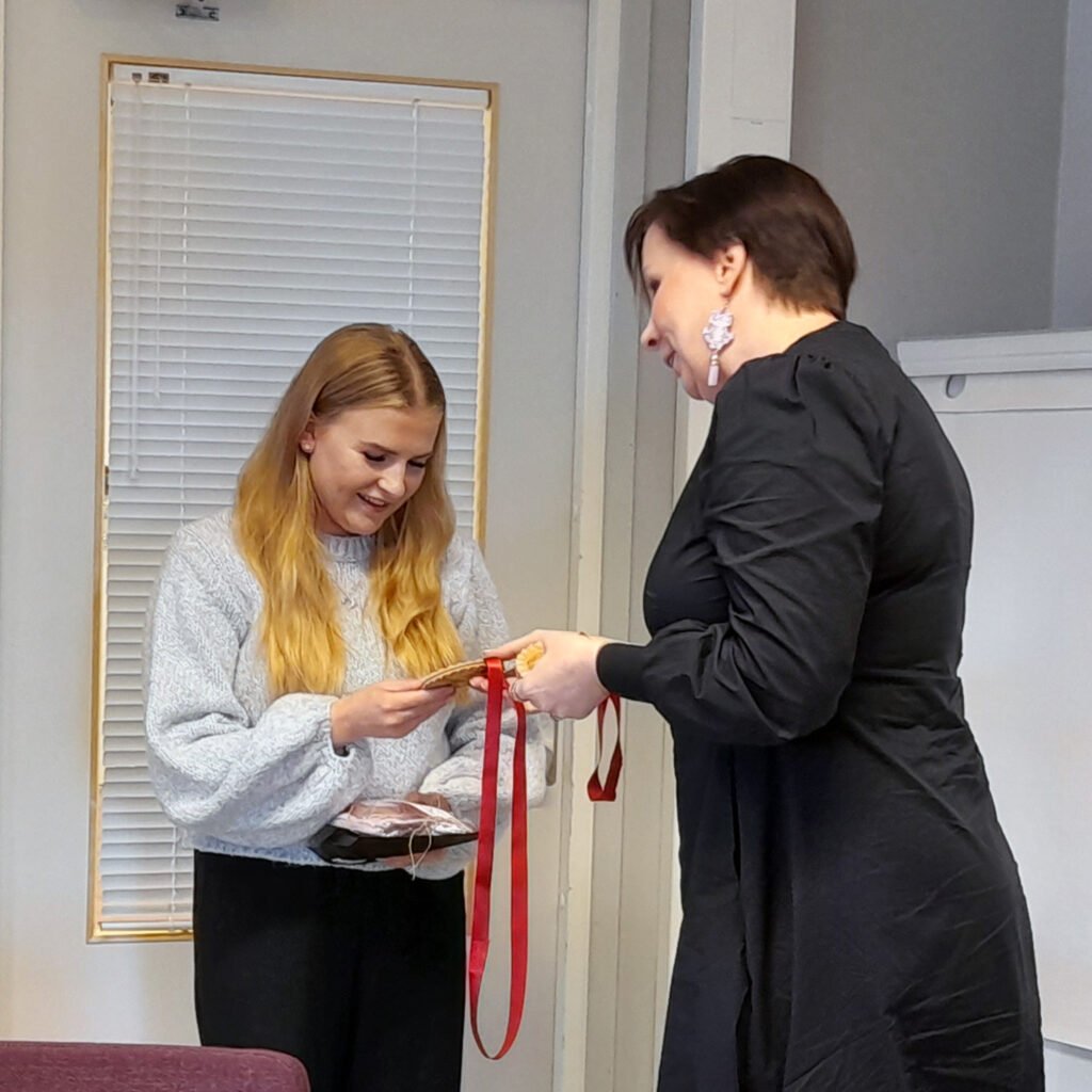 Nuori nainen vastaanottaa mitalin punaisella nauhalla vanhemmalta naiselta sisätiloissa. Molemmat hymyilevät tilanteessa.