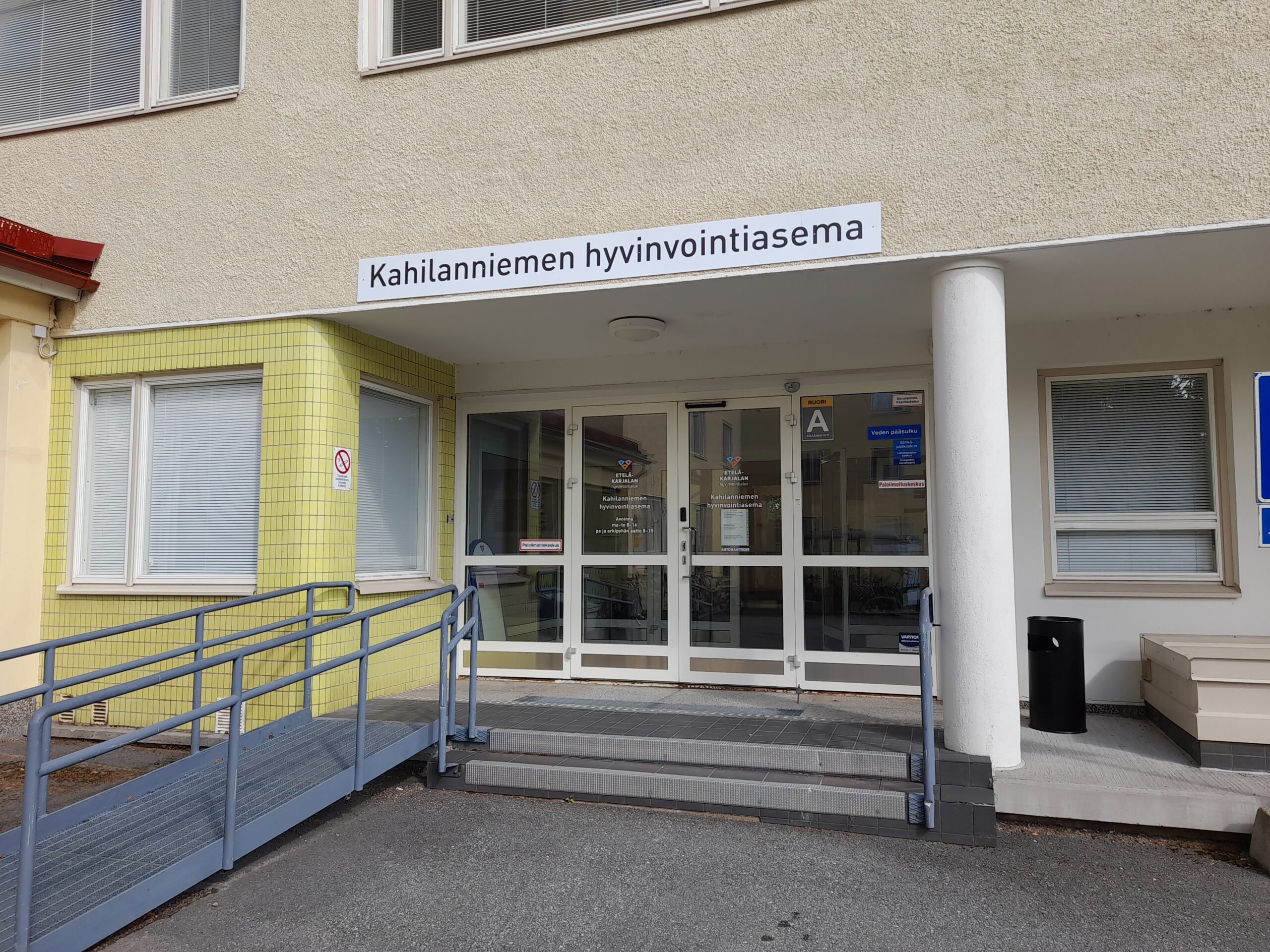 Rakennuksen sisäänkäynti, jossa on valkoinen kyltti tekstillä "Kahilanniemen hyvinvointiasema". Oveen johtaa sekä luiska että portaat. Ovien vieressä on ikkunoita, ja seinä on osittain vaaleankeltainen ja osittain vaaleanharmaa.