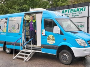 Sininen pakettiauto, jossa paljon kuvateippauksia. Auton sivuovi on avoinna ja auton sisällä seisoo sairaanhoitaja. Avonaiselle ovelle johtaa metallinen portaikko.
