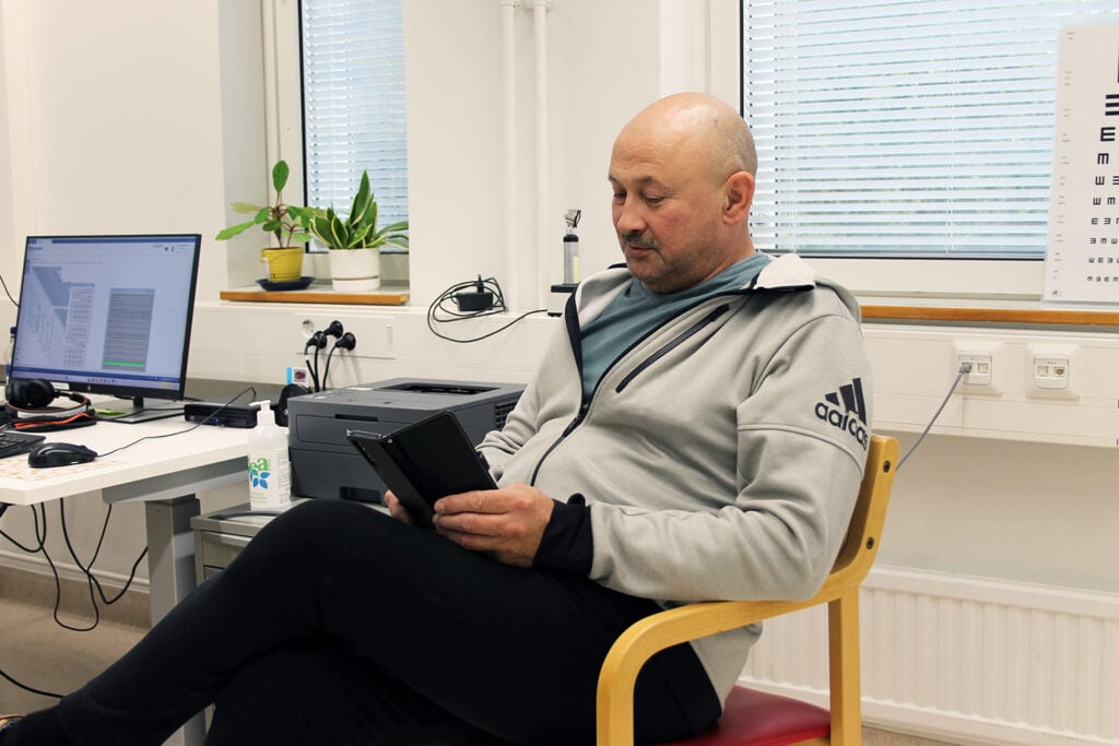 Henkilö istuu punaisella tuolilla vastaanottohuoneessa, pukeutuneena harmaaseen Adidas-huppariin ja mustiin housuihin. Henkilö pitää kädessään tablettia. Taustalla näkyy työpöytä, jossa on tietokoneen näyttö, näppäimistö, tulostin ja kasvi ikkunalaudalla. Ikkunassa on sälekaihtimet ja seinällä näkötaulu.
