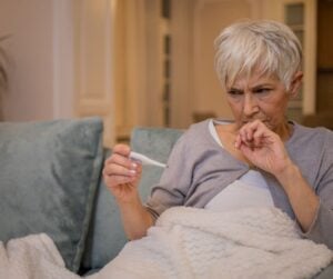 Harmaatukkainen nainen katsoo epäuskoisen näköisenä kädessään olevaa kuumemittaria. Nainen istuu sohvalla ja hänellä on vaalea peitto ympärillään.
