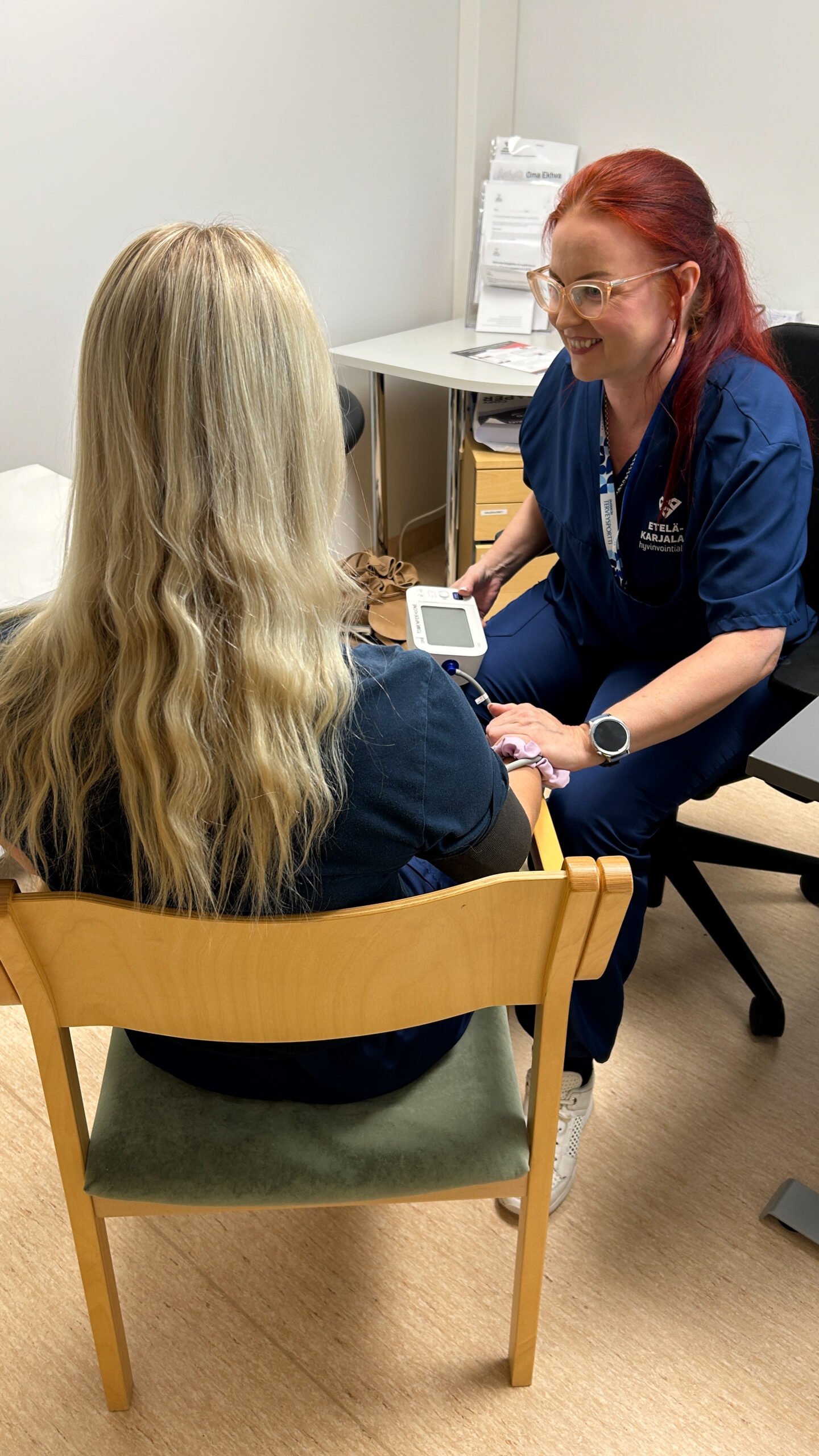 Milla mittaa verenpainetta asiakkaalta ja pitää kättä asiakkaan käden päällä. Milla hymyilee asiakkaalle.
