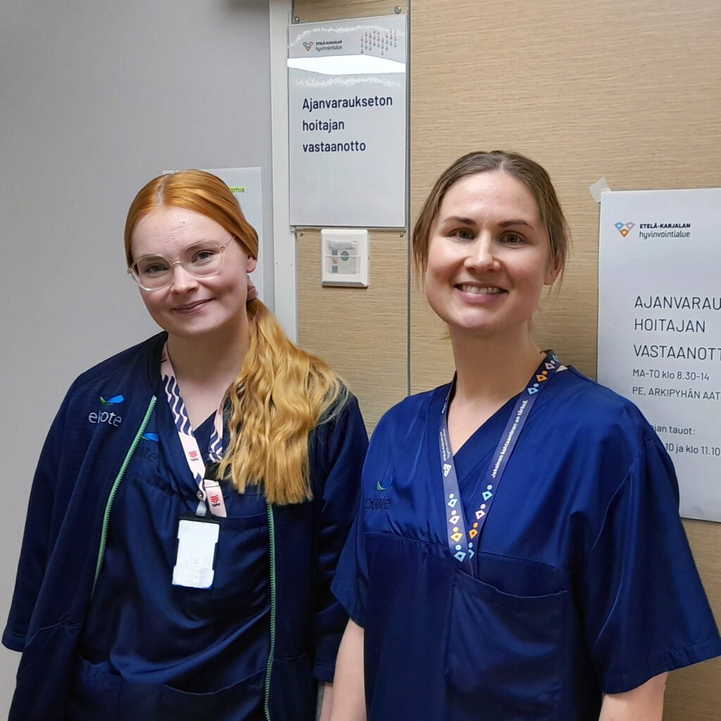 Kaksi hiukset taakse sitonutta tummansinisiin hoitajan asuihin pukeutunutta naista seisoo hymyillen hoitohuoneen oven edessä käytävällä.