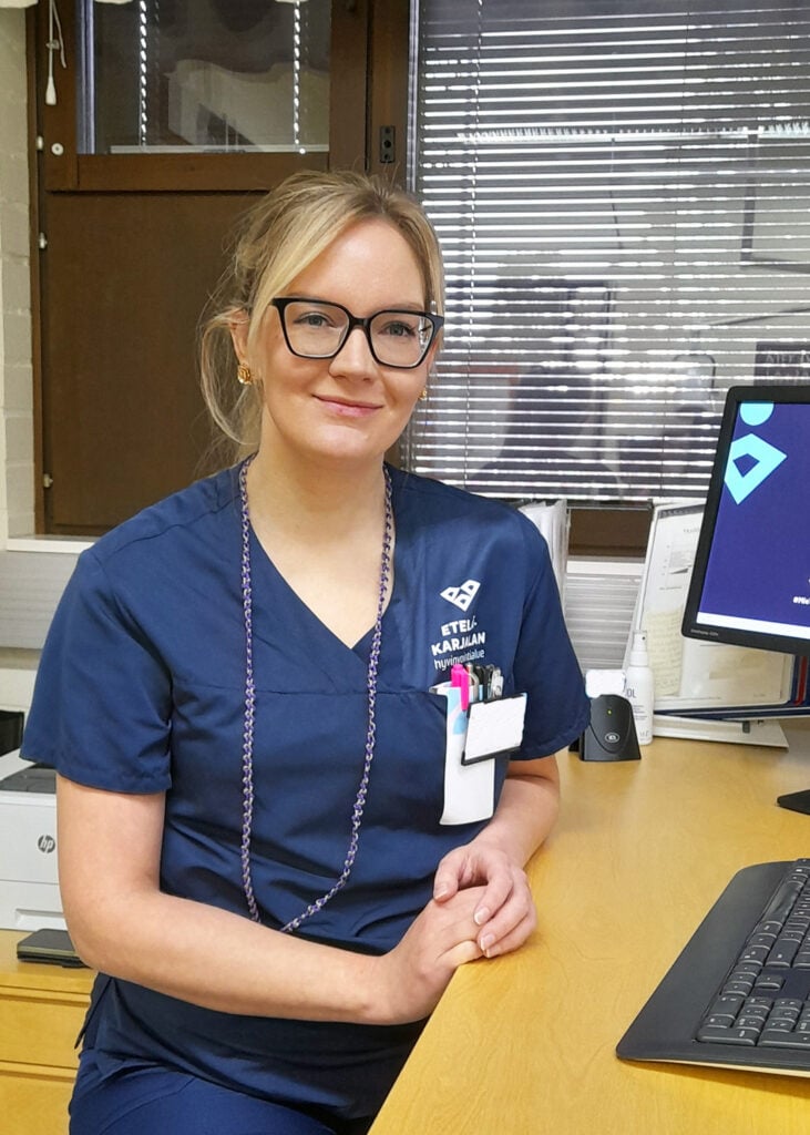 Sairaanhoitaja-asussa oleva nainen hymyilee ja katsoo kameraan työpöytänsä ääressä.