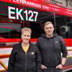 Hymyilevät nainen ja mies seisovat hälytysajoneuvon edessä. 