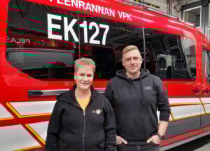 Hymyilevät nainen ja mies seisovat hälytysajoneuvon edessä.