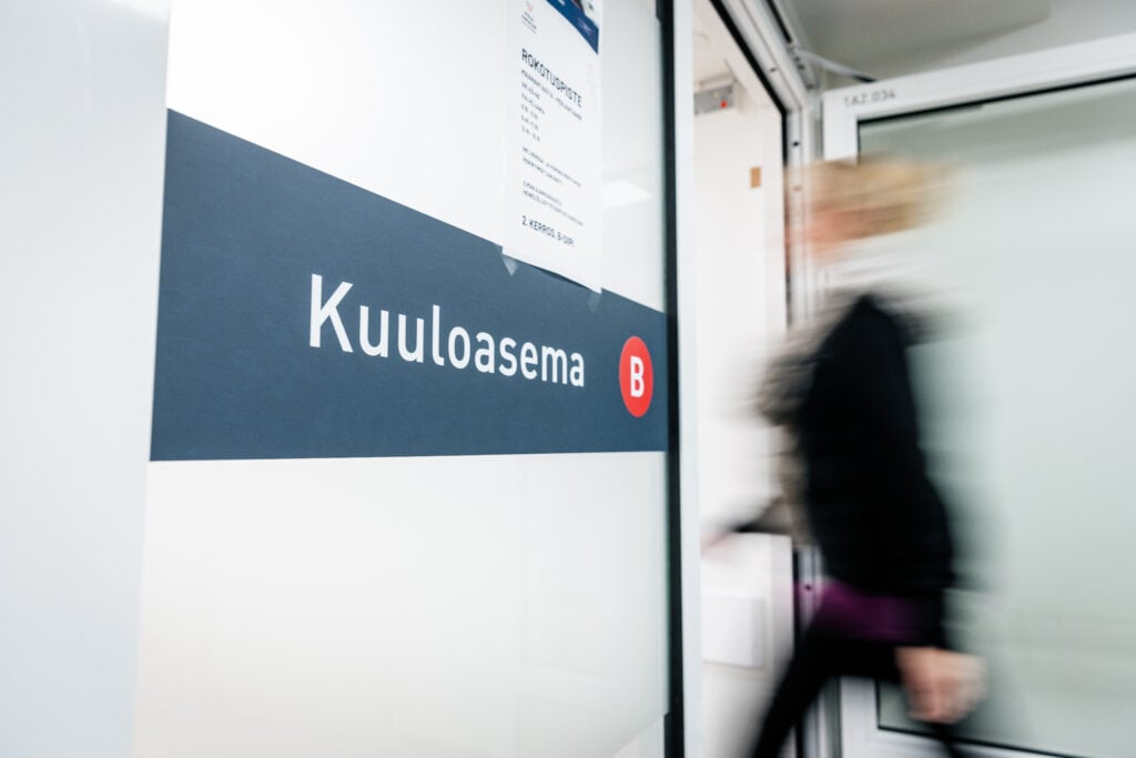 Opaste, jossa lukee kuuloasema. Taustalla henkilö astuu sisään viereisestä ovesta.