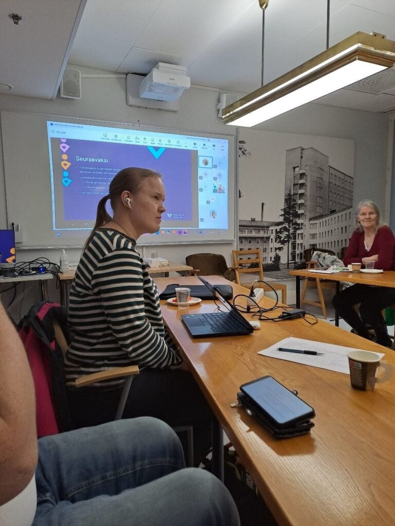 Kuvassa on henkilö sivuttain raidallisessa paidassa. Kuvassa iso neuvottelupöytä, jonka toisella puolella on punaiseen pukeutunut henkilö. Taustalla on seinälle heijastettu kuva, jossa diaesitys lilalla pohjalla.