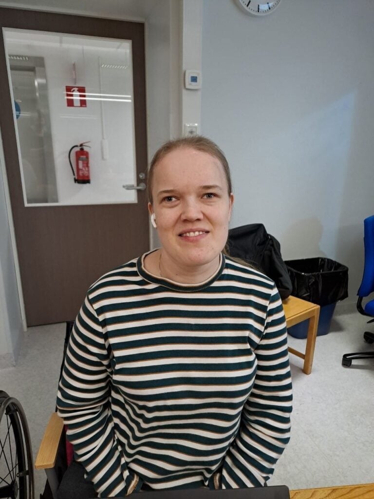 Kuvassa on sisätiloissa istuva henkilö, joka katsoo kameraan ja hymyilee kevyesti. Hänellä on yllään vihreä-valkoraidallinen pitkähihainen paita ja valkoiset nappikuulokkeet korvissa. Henkilö istuu pöydän ääressä, ja hänen takanaan näkyy ovi, jossa on palosammuttimen kuvake sekä seinään kiinnitetty palosammutin. 