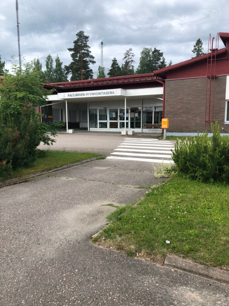 Rautjärjven hyvinvointiasemasta kuva, punatiilinen yksikerroksinen rakennus, jossa paljon valkoista puuta, etualalla sisäänkäynti piha-alueineen. 