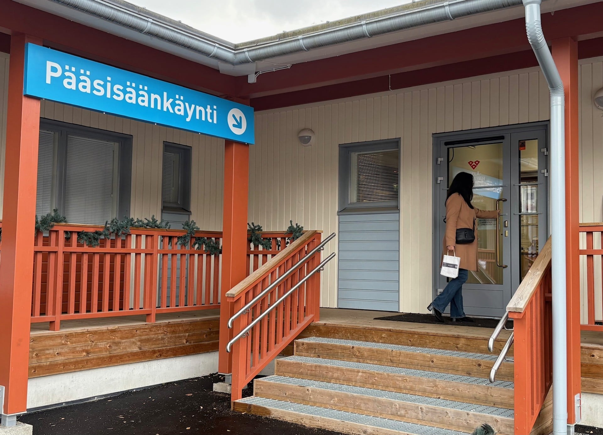 Hyvinvointiaseman portaiden yläpäässä nainen pitkässä takissa on avaamassa ovea. Nainen on selin kuvassa.