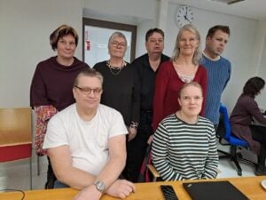 Kuvassa on ryhmä ihmisiä, joista 5 seisoo takana ja kaksi istuu edessä. Oikealla reunalla takana istuu yksi henkilö sivuttain. Taustalla on ovi ja seinällä kello.