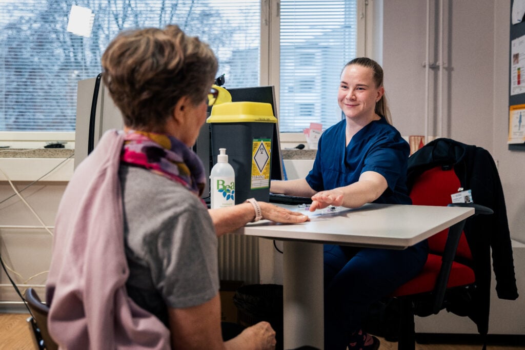 Hoitaja ja asiakas istuvat vastakkain pöydän ääressä terveydenhuollon vastaanottotilassa. Hoitaja tarkastelee asiakkaan kättä pöydällä. Pöydällä on käsihuuhdepullo ja taustalla näkyy ikkunat sekä hoitotarvikkeita.