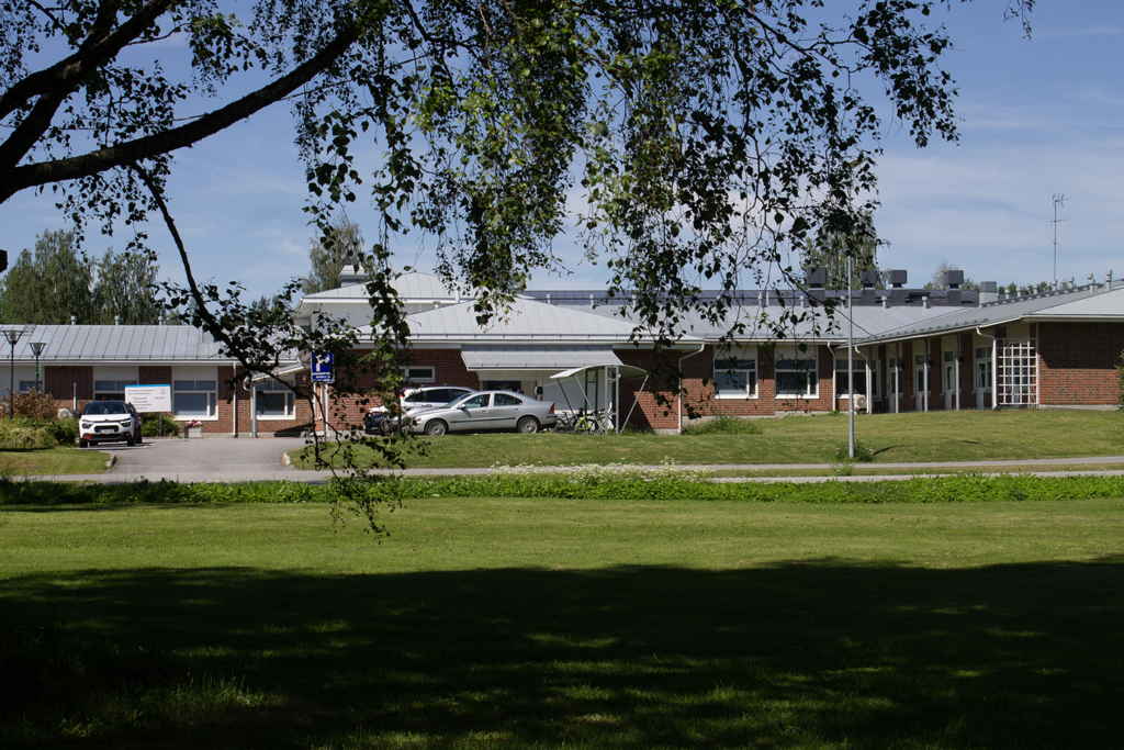 Kaukaa otettu ulkokuva matalasta punatiilisestä rakennuksesta. Etualalla vihreä nurmikkoalue. 