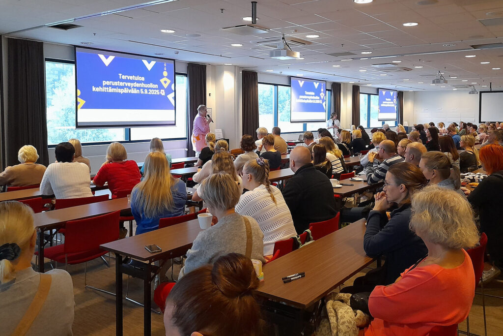 Takaapäin kuvattu suuri kokoustila, joka on aivan täysi istuvista ihmisistä. Edessä on nainen puhumassa mikkiin. Kolmelle isolle led-näytölle on heijastettu teksti Tervetuloa perusterveydenhuollon kehittämispäivään 5.9.2025.