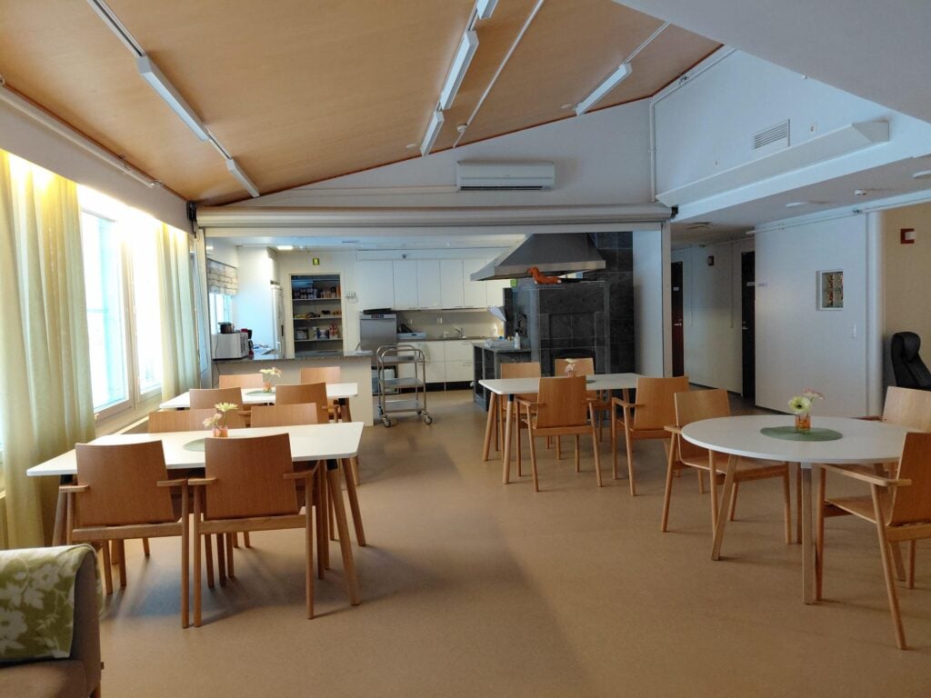 kuva keittiöstä, jossa pöytäryhmiä ja taustalla valkoinen keittiö. takana keittiön edustalla iso vuolukivinen leivinuuni. 