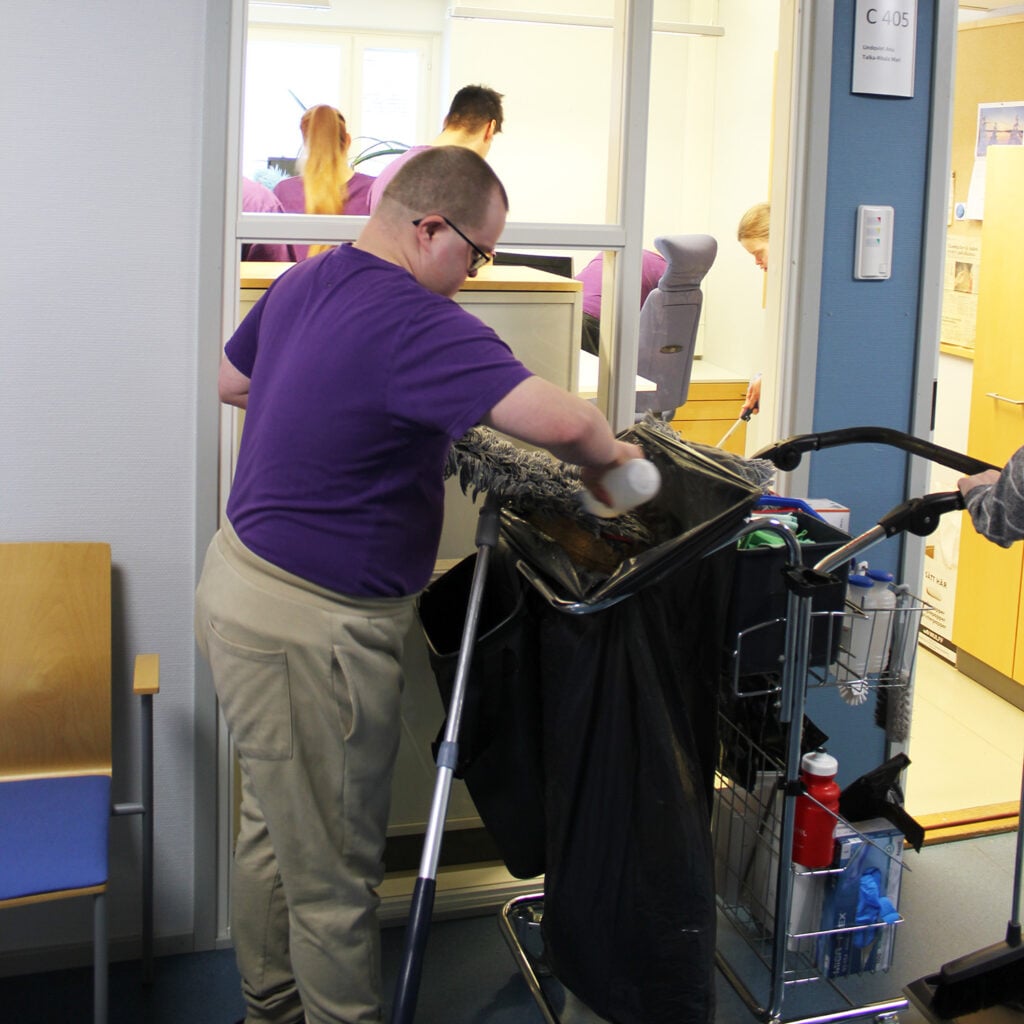 Puhtaanapidon työntekijä laittaa pesuaineitta moppiin, takana näkyy toimistossa muita siivoamassa