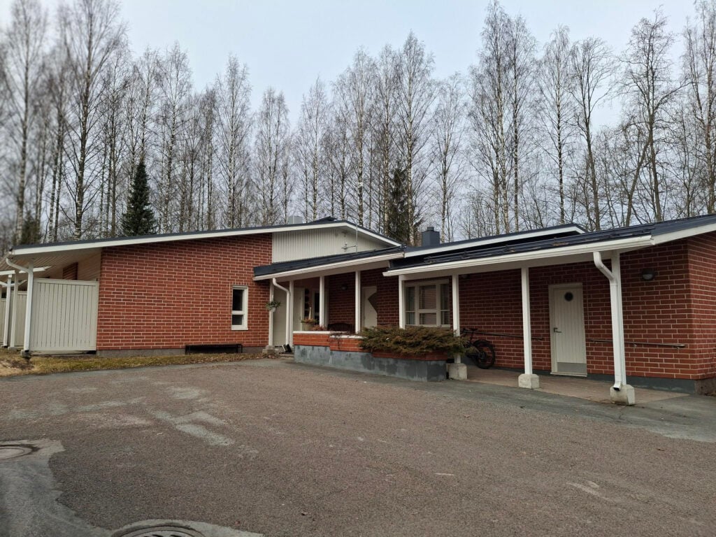 Punatiilinen matala rakennus, jossa on käytetty valkoisia puurakenteita. Etualalla on hiekkapintainen piha-alue.