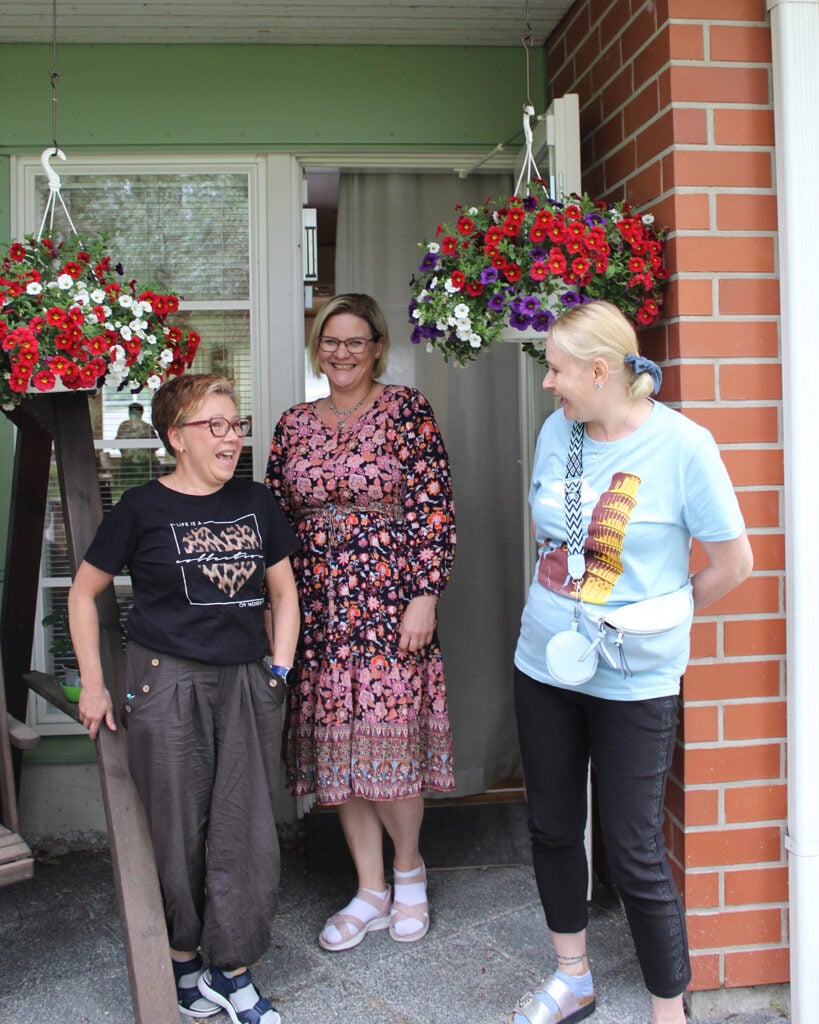 Kolme naista juttelevat keskenään ja nauravat, taustalla näkyy kaksi isoa kukka-amppelia