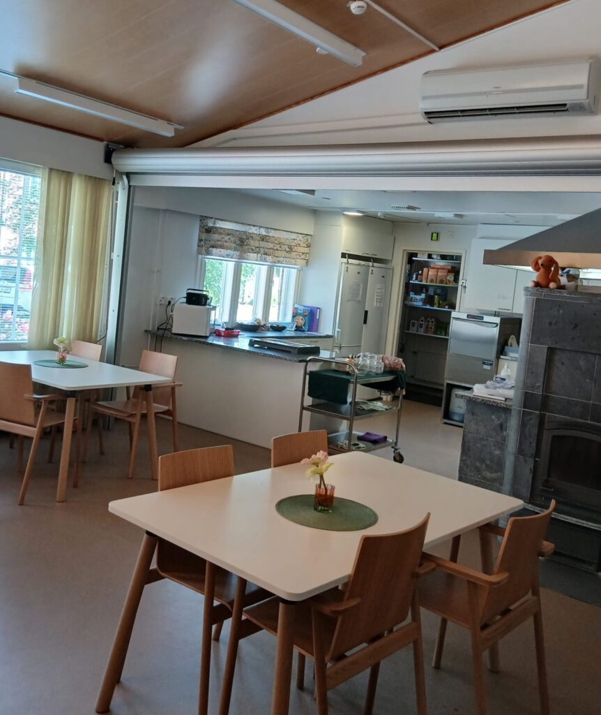 Sydänrinteen ruokasali ja keittiö. Pöytiä, vuolukiviuuni ja tiskin takana keittiövarustusta.