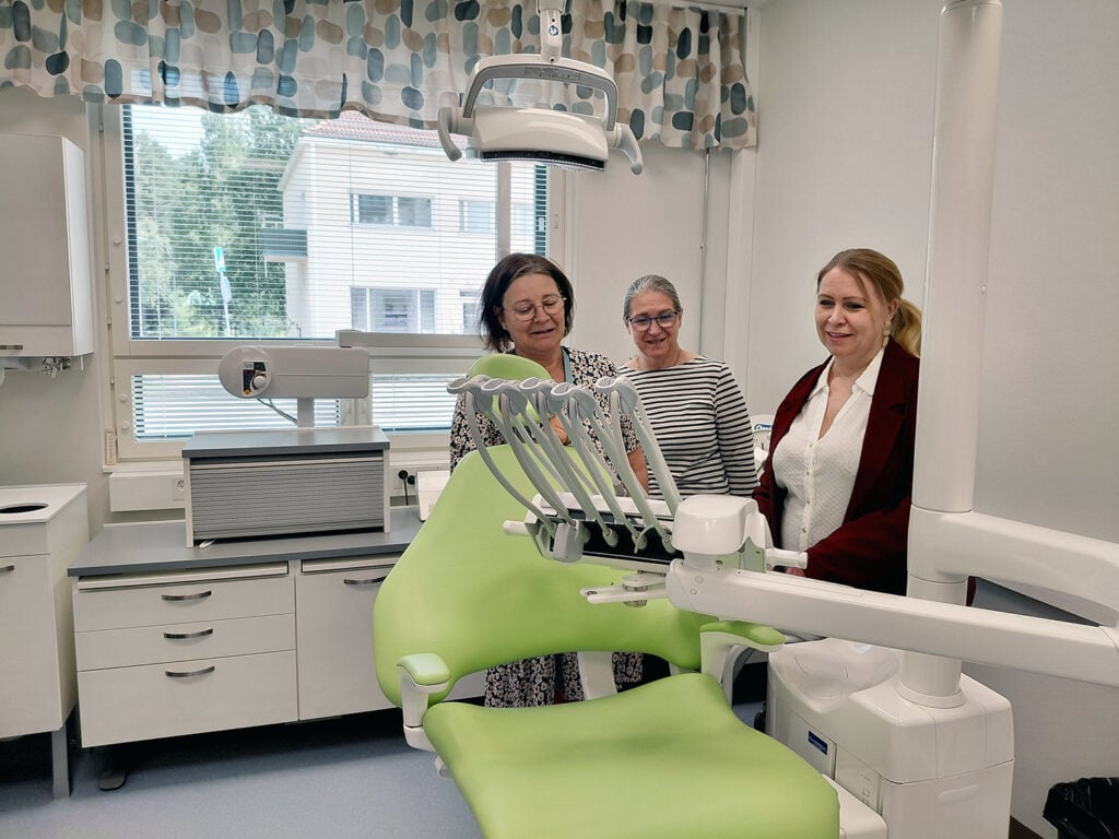 Valokuva hammashoitohuoneesta, jossa limen värinen hoitotuoli ja sen takana kolme hymyilevää naista. 