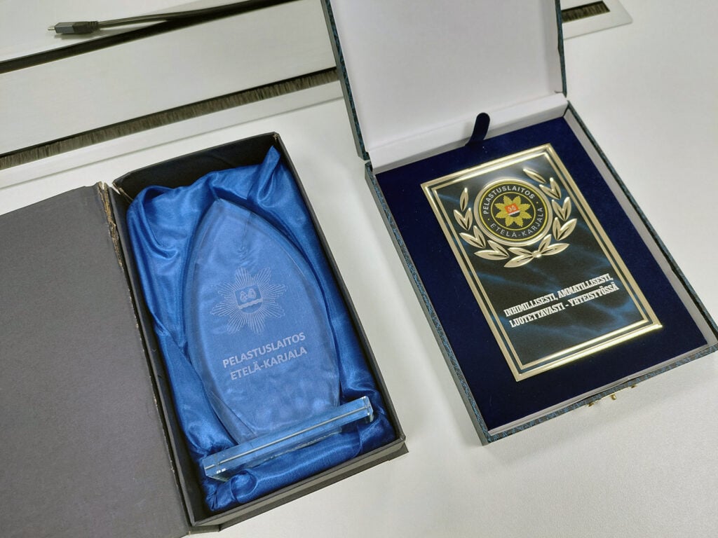 Lähikuva Elämän liekki -palkinnoista. Toinen on lasinen liekki sinisellä kankaalla vuoratussa pahvikotelossa ja toinen on metallisen oloinen laatta, jossa teksti Elämän liekki, Inhimillisesti, ammatillisesti, luotettavasti, yhteistyössä.