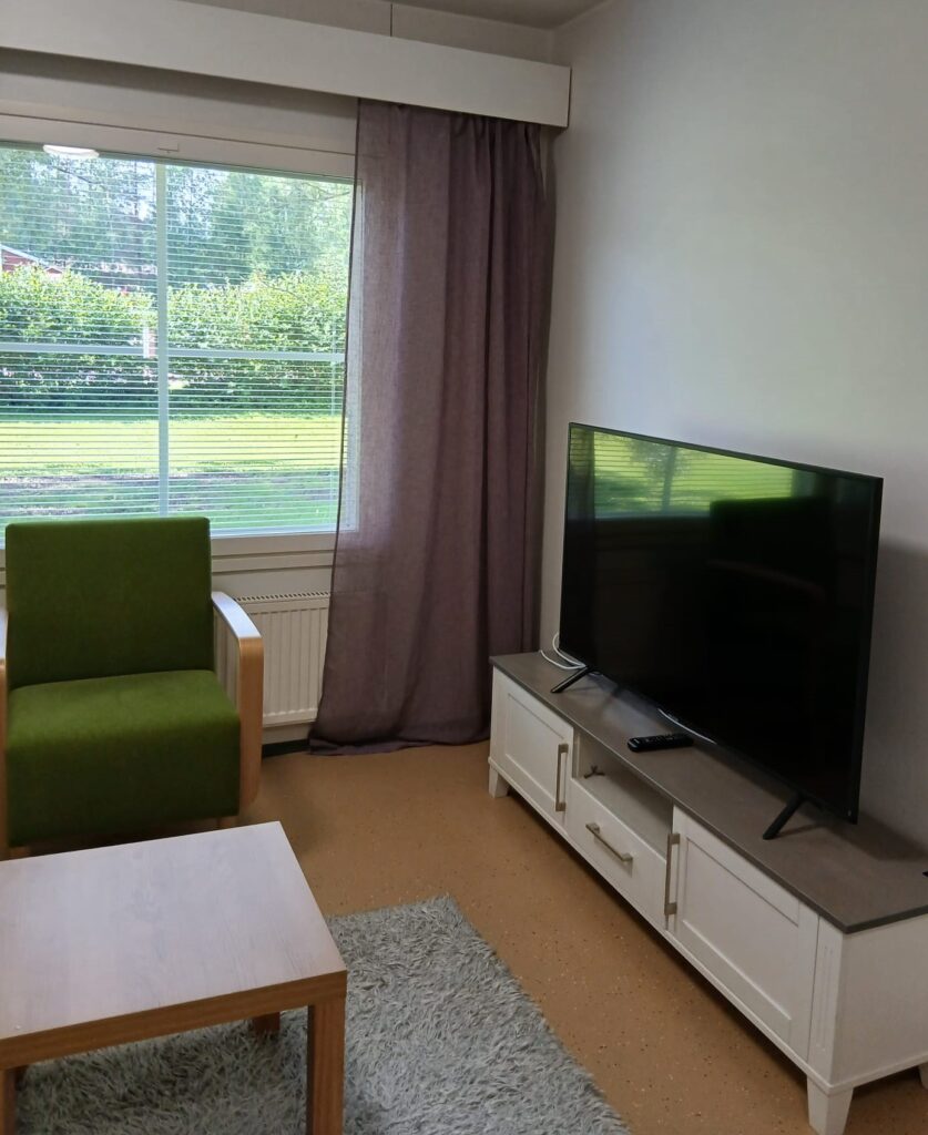 Huone, jossa on nojatuoli, pöytä ja lipaston päällä televisio.